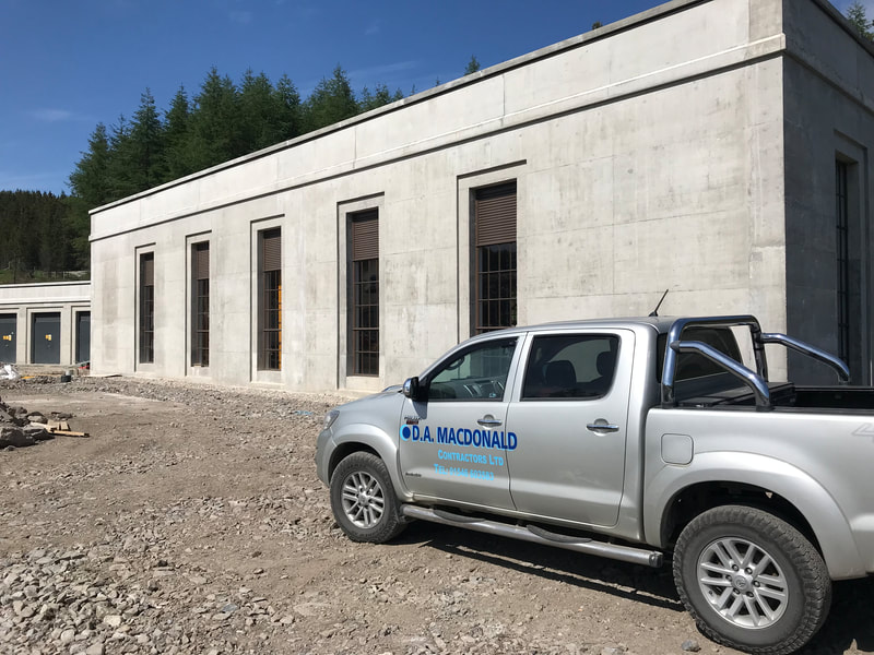 A concrete power station building with tall brown windows, and a silver 4x4 in the foreground